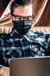 © Ekaterina - Remote operation during the quarantine, to print the text and conduct important business. A handsome young freelancer with diopter glasses and a black protective mask works on a silver laptop at home.