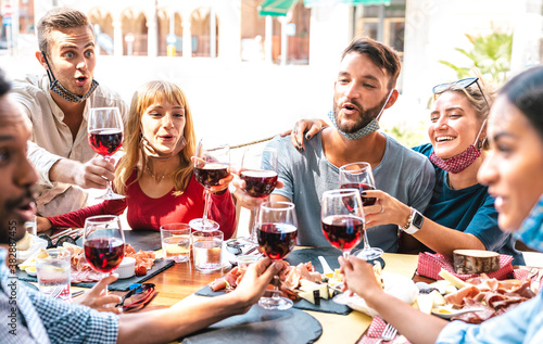 Friends toasting red wine at restaurant bar wearing opened face masks - New normal lifestyle concept with happy people having fun together on sunny day - Bright filter with focus on left blond woman