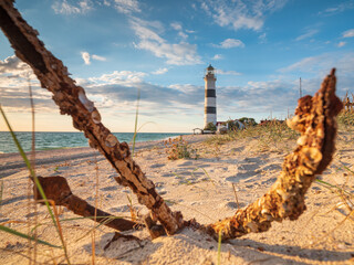 Wall Mural - close up view to old metal anchor on the sand with wide angle view to lighthouse under blue sky in sunset
