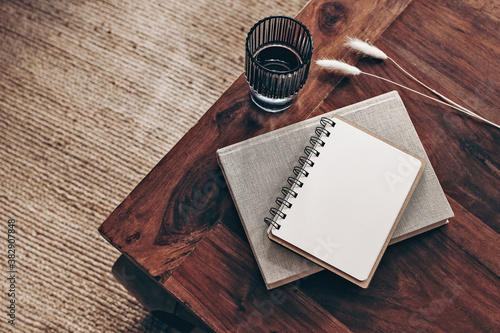 Empty diary, notepad mockup and book on wooden table. Glass of water and dry Lagurus ovatus grass. Blurred beige jute carpet background. Flat lay, top view. Simple Scandinavian boho interior.