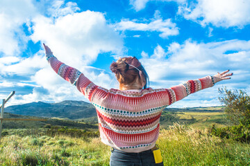 Young woman with open arms and colorful clothes style looking at a natural mountains and grass with a big blue sky. Colorful and emotional landscape