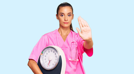 Wall Mural - Young hispanic woman as nutritionist doctor holding weighing machine with open hand doing stop sign with serious and confident expression, defense gesture