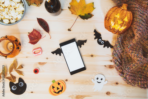 Top view smart phone with blank screen decorated with bat wings and traditional Halloween decor - bats, ghost silhouettes, lighting pumpkin, fall leaves, sweets, warm plaid on the wooden background.