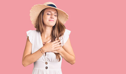 Young beautiful blonde woman wearing summer dress and hat smiling with hands on chest with closed eyes and grateful gesture on face. health concept.