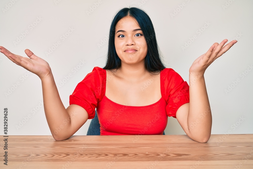 Young beautiful asian girl wearing casual clothes sitting on the table clueless and confused expression with arms and hands raised. doubt concept.