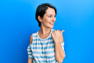 Young brunette woman with short hair wearing casual striped t-shirt smiling with happy face looking and pointing to the side with thumb up.