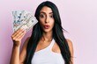 © Krakenimages.com - Beautiful hispanic woman holding dollars scared and amazed with open mouth for surprise, disbelief face