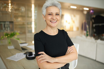 Portrait of joyful confident gray haired mature female freelancer in casual clothes smiling broadly at camera having break while working from home, posing against modern interior background