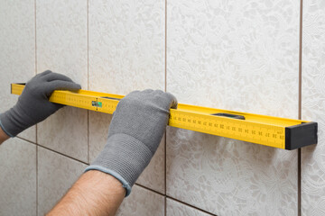 Worker hands in protective gloves measuring tiles surface on wall with yellow spirit level. Checking result after tiles gluing. Precision work. Closeup.