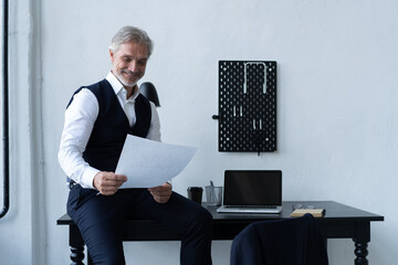 Pensive handsome matured male owner of corporation reading financial documentation standing near table, confident businessman in trendy formal wear checking accounting report