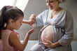 © Halfpoint - Small girl creaming belly of pregnant mother indoors in bathroom at home.