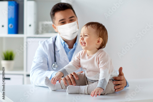 Fototapeta medicine, healtcare, pediatry and people concept - happy doctor or pediatrician wearing face protective mask for protection from virus disease with baby on medical exam at clinic
