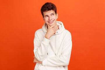 Wall Mural - Smiling young man with peaceful face thinking over plans for future holding his chin, thoughtful teenager dreaming about good job. Indoor studio shot isolated on orange background