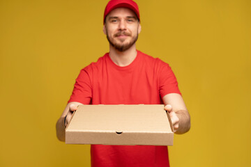 Wall Mural - Photo of happy man from delivery service in red t-shirt and cap giving food order and holding pizza box isolated over yellow background