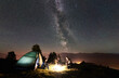 Young romantic couple hikers having a rest at campfire beside camp and tourist tent under night sky full of stars and Milky way. On the background beautiful starry sky, mountains and luminous town
