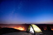 Summer camping on the top of mountain in the evening. Glowing tent under amazing night sky full of stars. On the background incredibly beautiful starry sky, mountains and luminous city
