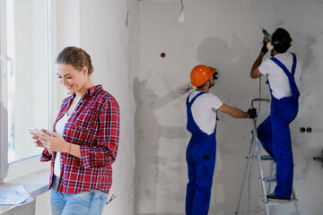 A woman in a plaid shirt is texting by phone. Two general laborer in helmets and headphones are drilling the wall