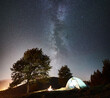 Tourist camping near forest at night. Illuminated tent and bonfire under amazing night sky full of stars and Milky way. On the background beautiful starry sky, mountains and luminous town