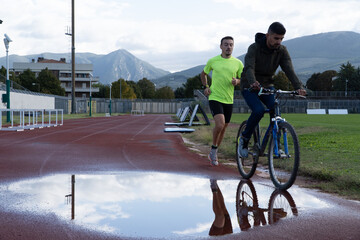 Two men training on a training track. One is running and the other is cycling.