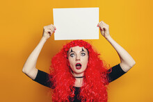 A Girl In A Halloween Costume Holds A Sheet Of Paper Above Her Head. Young Shocked Woman In Red Curly Wig Holding White Sheet Of Paper And Looking At It Isolated On Yellow Background
