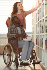 Wall Mural - Vertical full length portrait of young African-American man using wheelchair in school with female friend helping him in library lit by sunlight