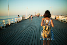 Young Female Tourist With Vintage Backpack Walking On The Wooden Pier In Golden Hour