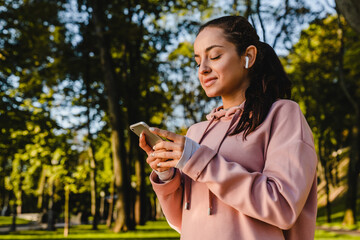 Smiling sporty girl is using her phone with earbuds walking in a park