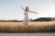Beautiful young blonde woman with long hair on a summer afternoon jumping high in the field. Happy girl in white dress looks at camera smiling