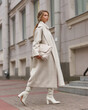 Elegant woman in white dress, hessian boots and coat waling at city street. Fall autumn fashion look. Pretty tall stylish young gitl with fashionable makeup and hair style. Elegant lady. Full length