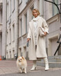Elegant woman in white dress, hessian boots and coat waling at city street. Fall autumn fashion look. Pretty tall stylish young gitl with fashionable makeup and hair style. Lady with german spitz