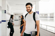 © kleberpicui - Portrait of young Brazilian student with backpack carrying books in college. Other students in the background.