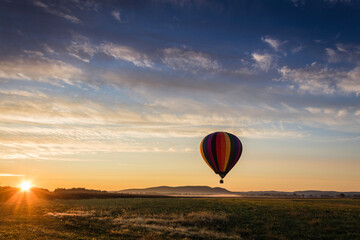 Wall Mural - Hot Air Balloon in colorful rainbow stripes begins ascent over farm field as sun rises blue cloudy sky