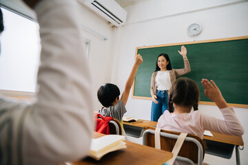 Wall Mural - Asian school teacher with students raising hands. Young woman working in school with arm raised, school children putting their hands up to answer question, enthusiasm, eager, enjoyment.