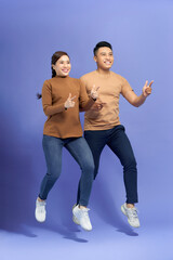 Wall Mural - Energetic beautiful couple posing together on camera while running or jumping and pointing fingers on copyspace along background