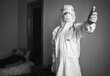 Black and white image of doctor in PPE suit, respirator, gloves and phonendoscope, holding contactless infrared thermometer, visiting patient at home during quarantine. Covid 19 concept