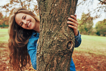 Wall Mural - Happy young woman smiling, wearing a blue denim shirt hugging a big tree, posing on nature background. Gorgeous female playing outdoor in the park. 