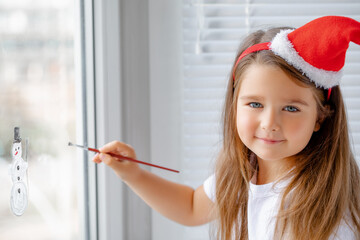 Wall Mural - Child Draws on Window on Christmas eve . Little Girl in a Santa Claus hat paints with colored paints Christmas Snowflakes and a Snowman, Decorates the house 