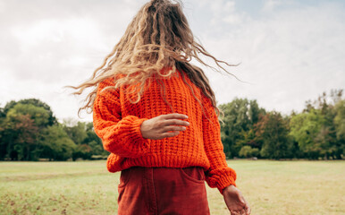 Horizontal image of an young woman with blowing long blonde hair wearing an orange sweater posing on the sky and nature background. Pretty female playing with her hair outdoor in the park. 