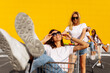 © Shopping King Louie - Two beautiful happy young women, girlfriends have fun on shopping carts, against the background of a yellow wall of a shopping center on a summer sunny day