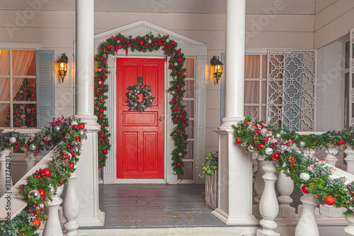 Christmas porch decoration idea. House entrance with red door decorated for holidays. Red and green wreath garland of fir tree branches and lights on railing. Christmas eve at home.