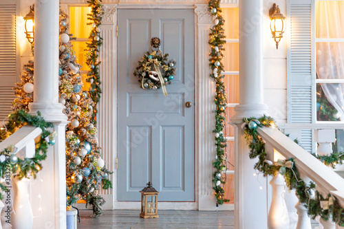 Christmas porch decoration idea. House entrance decorated for holidays. Golden and green wreath garland of fir tree branches and lights on railing. Christmas eve at home.