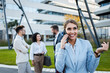 © Zoran Zeremski - Portrait of smiling businesswoman talking at smartphone while standing in front of modern office buildings with colleagues behind her.