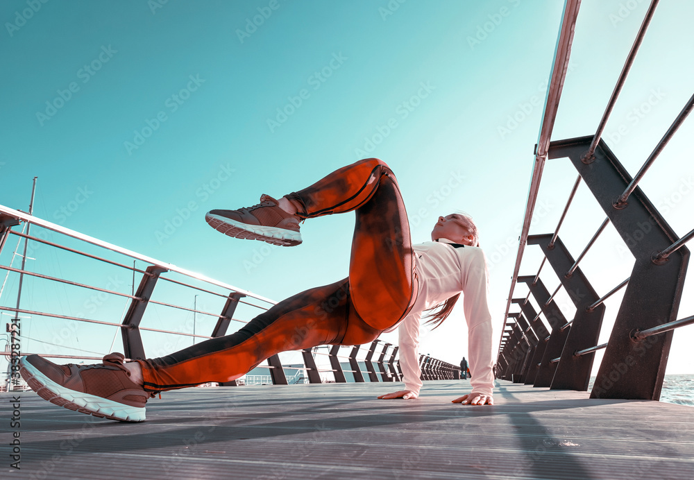 outdoor workout concept. young woman in tracksuit performs sports workout on the pier - obrazy, fototapety, plakaty 