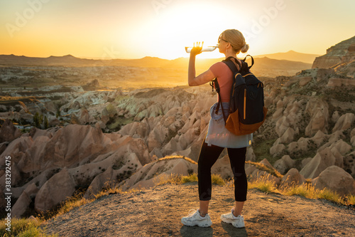 Woman backpacker drinking water at top of the mountain with breathtaking view on the valley during sunset