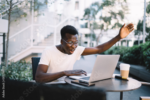 Angry dark skinned man in eyewear screaming while using laptop computer for remote freelance job, emotional dark skinned male freelancer stressed with software problem sitting on cafe terrace