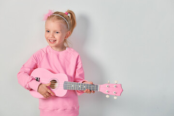 cute caucasian little girl 4-5 year old in pink sweatshirt are playing the ukulele against gray stud