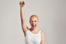 Portrait Of A Beautiful Young Caucasian Woman With Shaved Head Wearing White Shirt, Winking At Camera And Raising Clenched Fist, Standing Isolated Over Grey Background