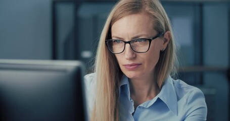 Close up of concentrated blonde in blue shirt and glasses sitting at table and using modern computer. Mature business woman working at bright office center.