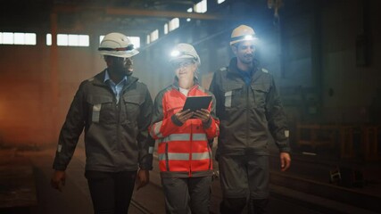 Wall Mural - Three Diverse Multicultural Heavy Industry Engineers and Workers in Uniform Walk in Dark Steel Factory Using Flashlights on Their Hard Hats. Female Industrial Contractor is Using a Tablet Computer.