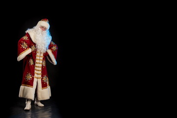 Santa Claus in a red fur coat, with a white beard and a bag of gifts posing on a black background